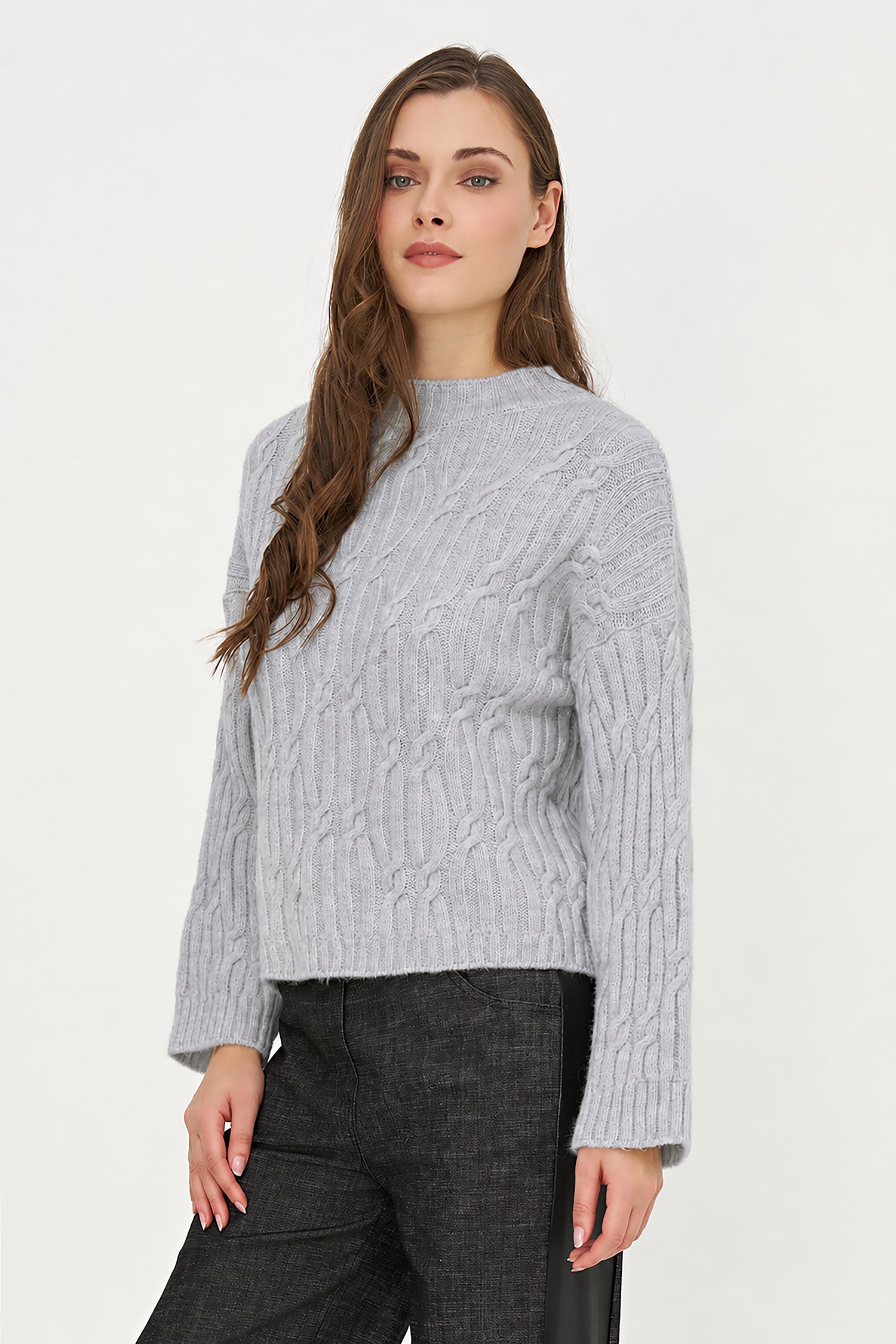A woman wearing a light gray cable-knit mock neck sweater and dark gray trousers, standing against a plain white studio background.