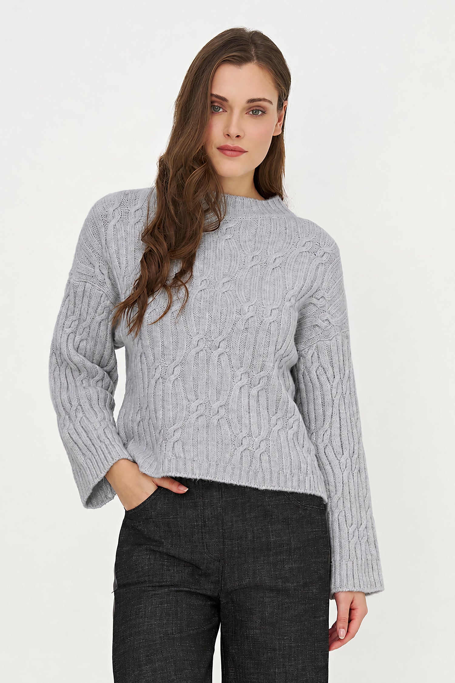 A woman wearing a light gray cable-knit mock neck sweater and dark gray pants, standing against a plain white studio background.