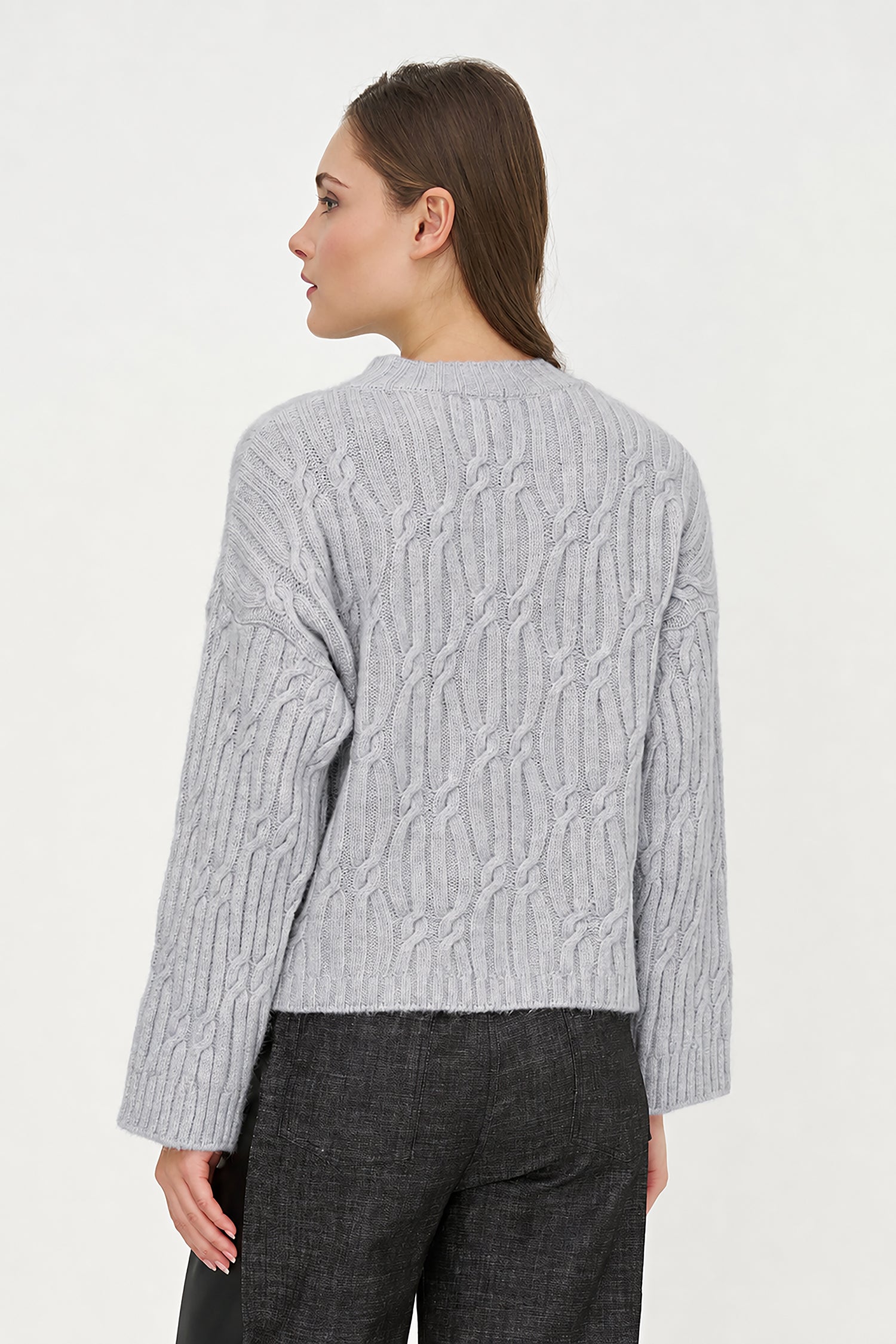 A woman shown from the back wearing a light gray cable-knit sweater and dark gray trousers, photographed against a plain light background.