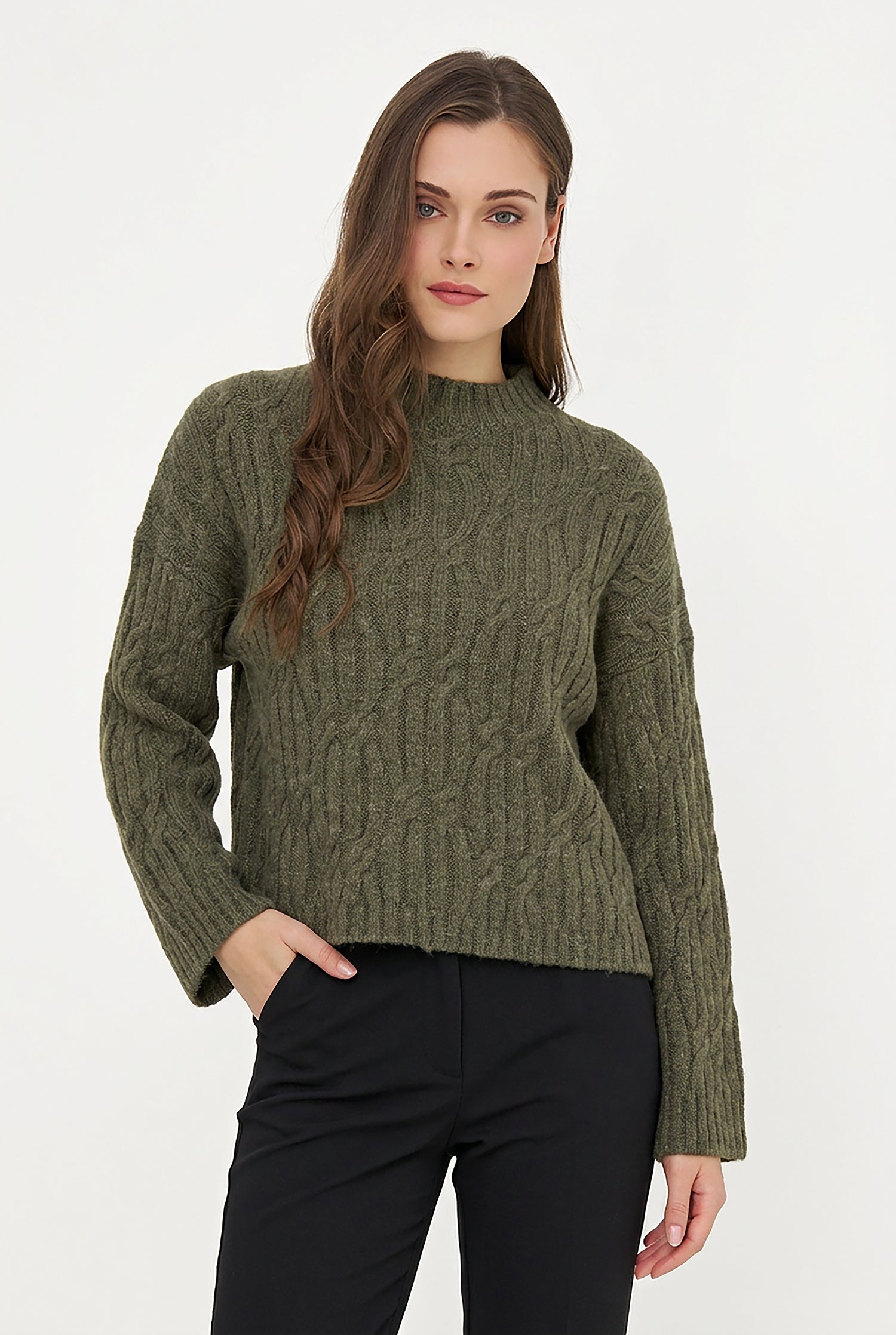 A woman wearing a textured olive green cable-knit mock neck sweater and black pants, standing against a plain white studio background.
