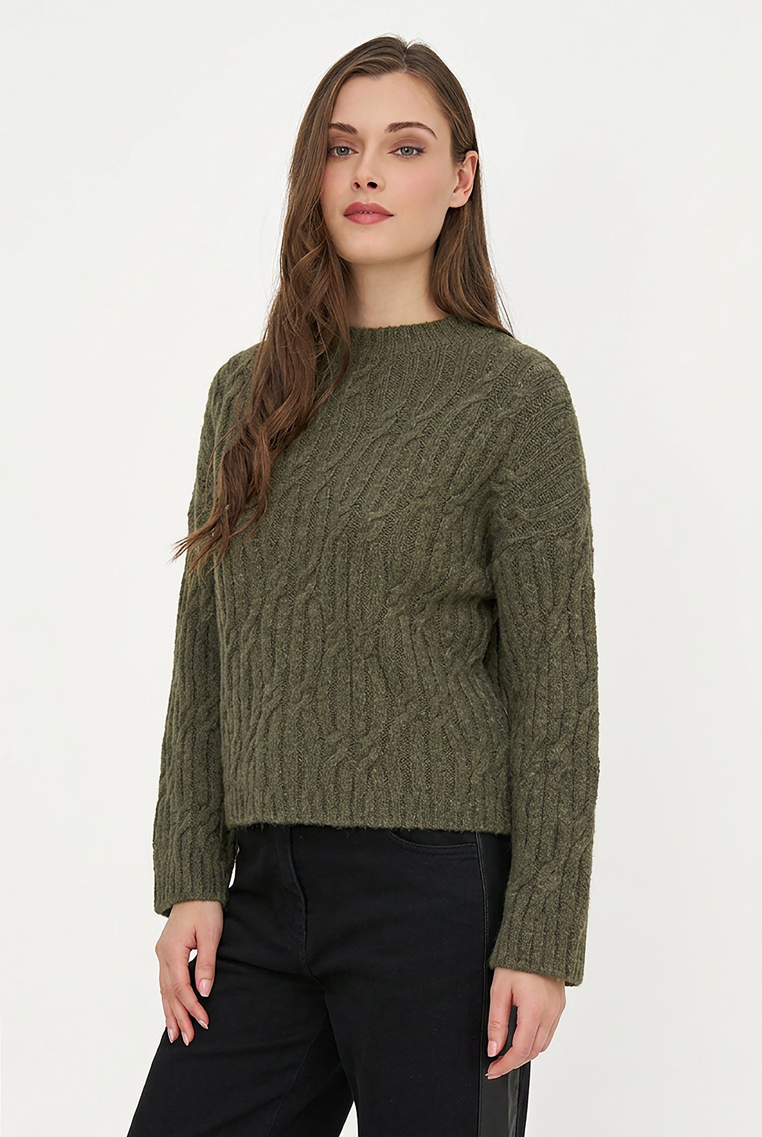 A woman wearing a textured olive green cable-knit sweater and black pants, standing against a plain white studio background.