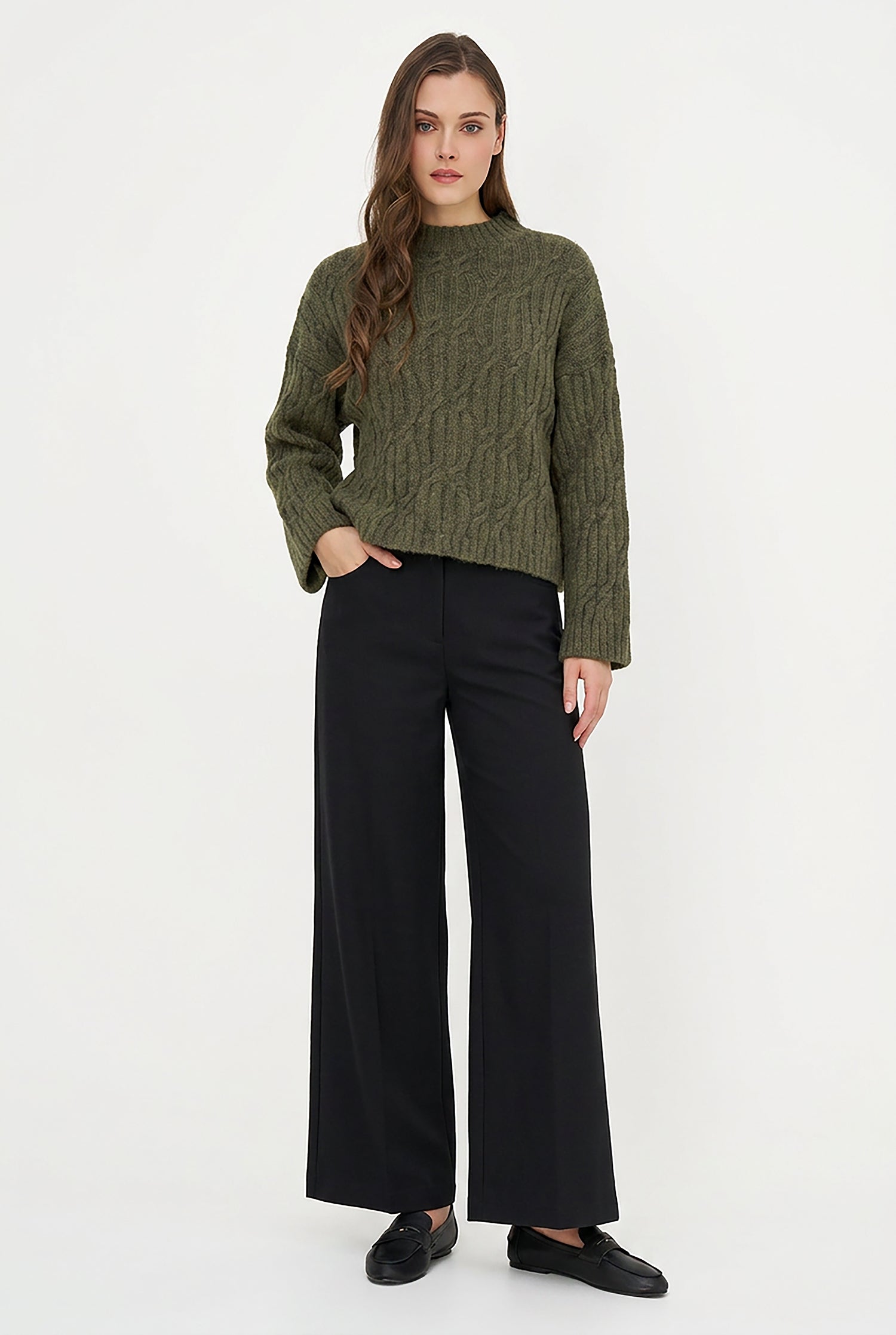 A woman modeling a cropped, olive green cable-knit sweater paired with black wide-leg trousers and black flat shoes, standing against a plain white studio background.