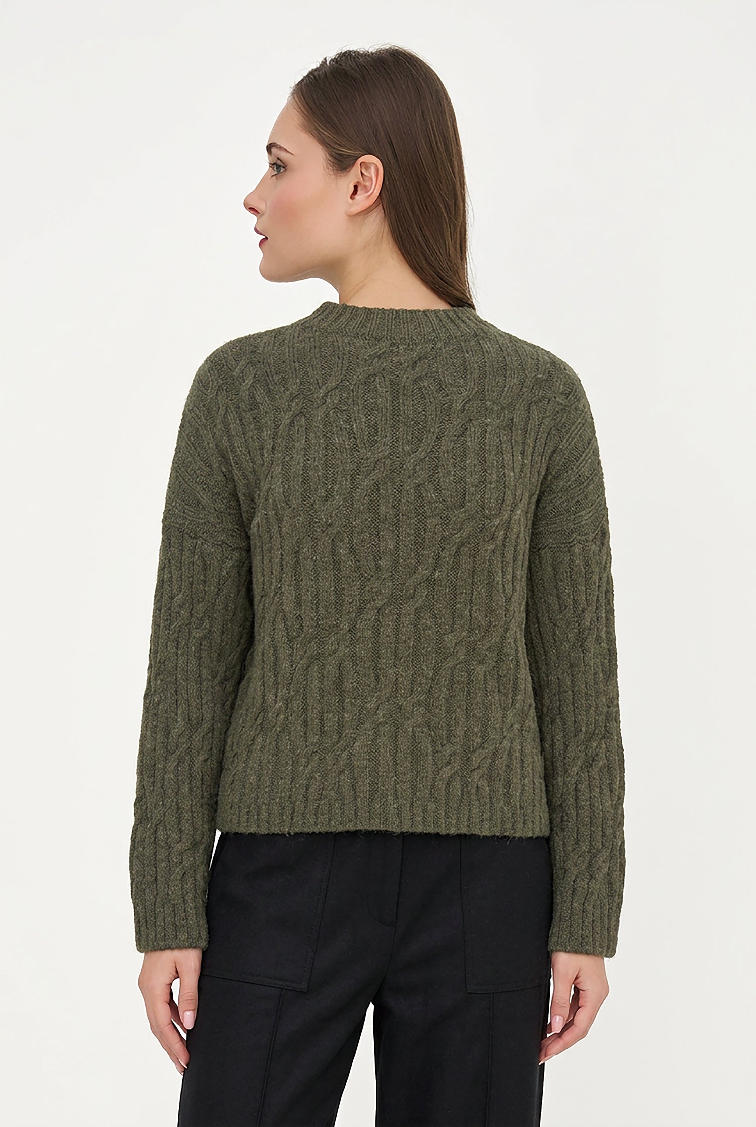 A woman shown from the back wearing an olive green cable-knit sweater and black trousers against a plain white studio background.