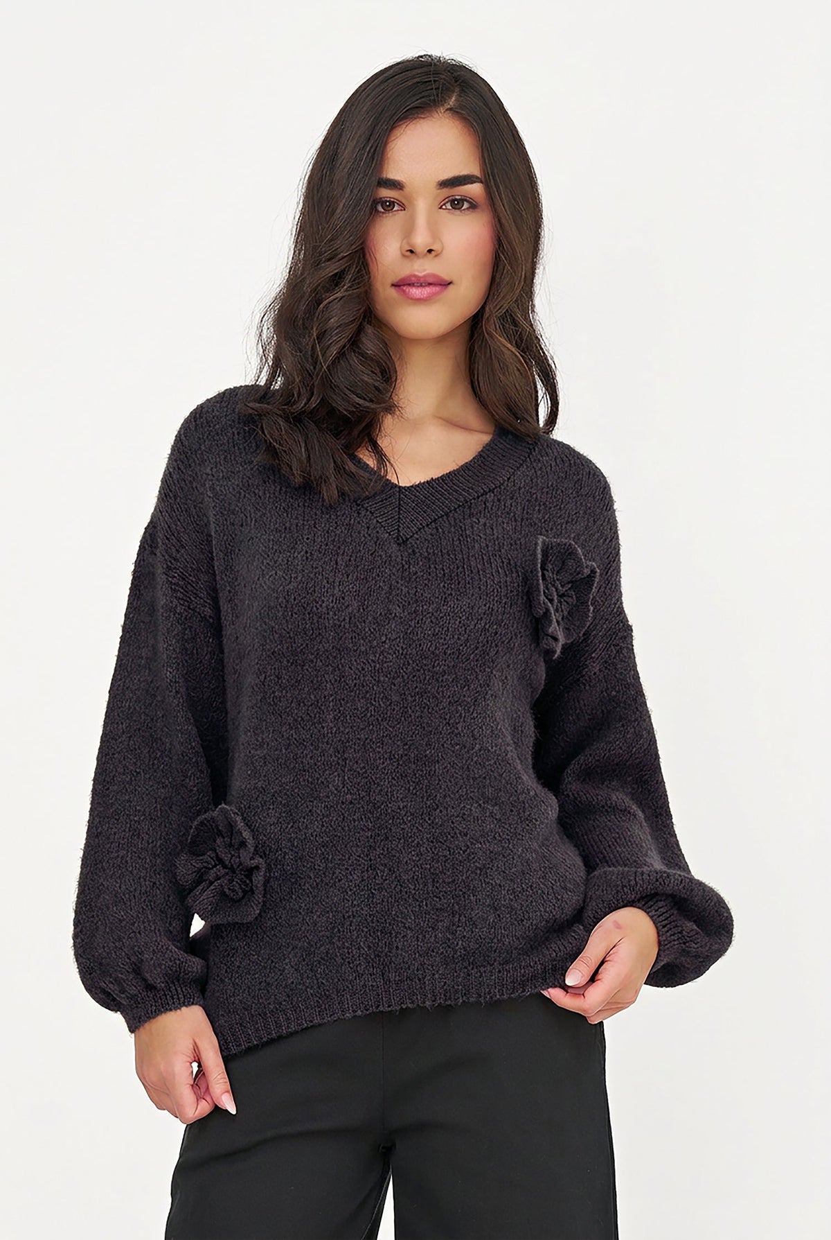 A woman wearing a dark charcoal gray fuzzy knit sweater with a large matching fabric flower detail on the chest, paired with black pants, against a plain white studio background.