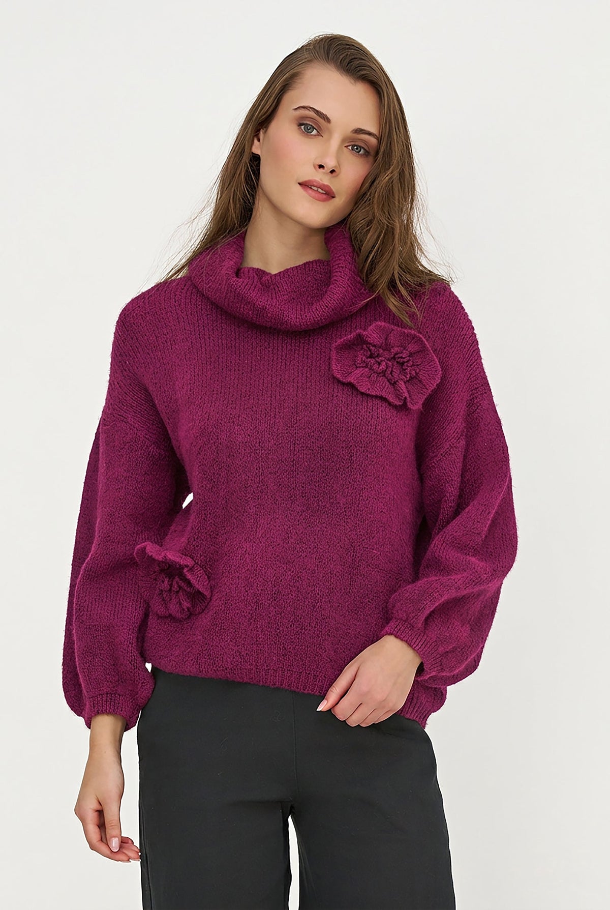 A woman wearing a textured magenta cowl-neck sweater with large fabric flower details on the chest and sleeve, paired with black trousers, against a plain light gray background.