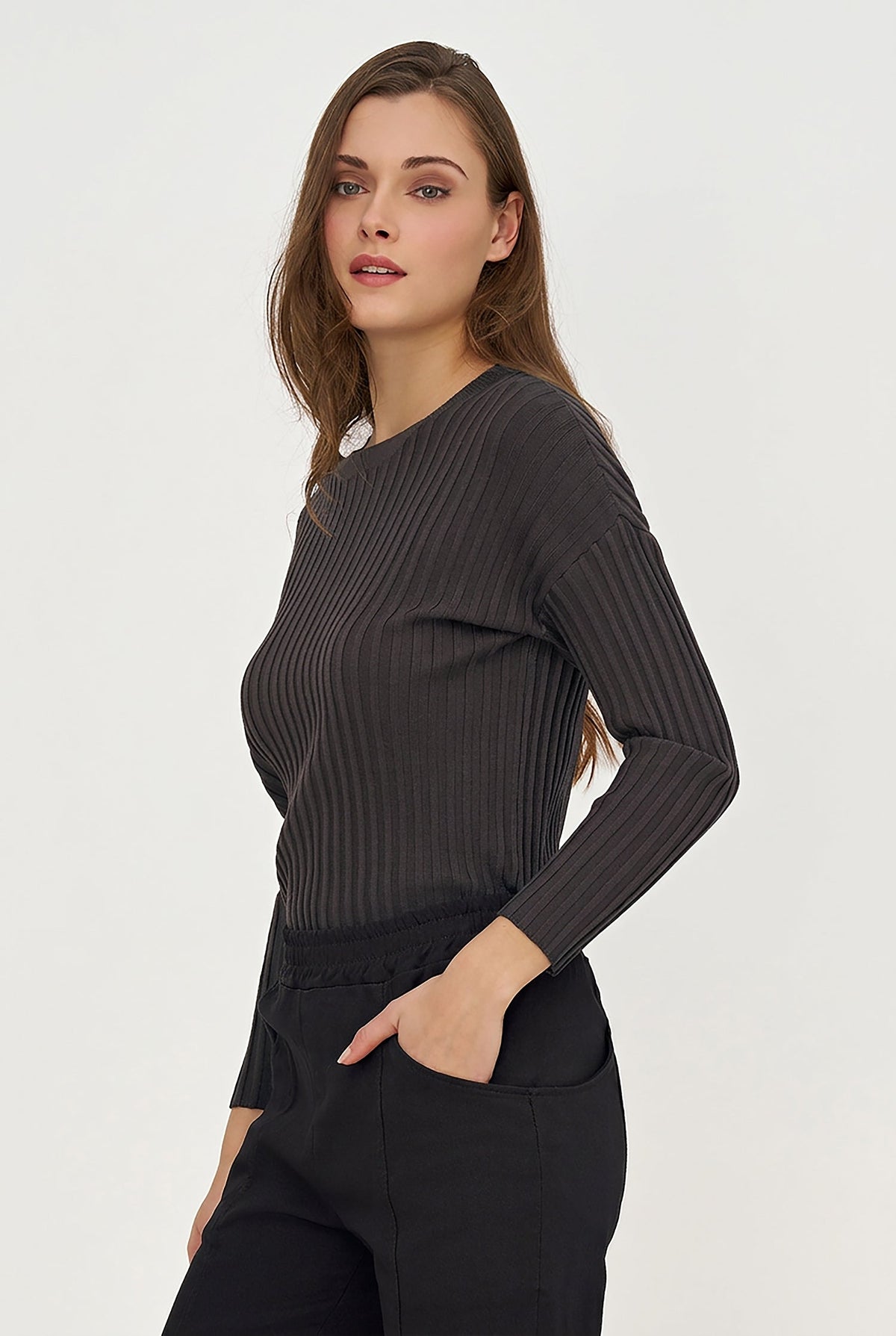 A woman modeling a dark charcoal gray ribbed knit long-sleeve top with black pants, photographed against a clean off-white studio background.