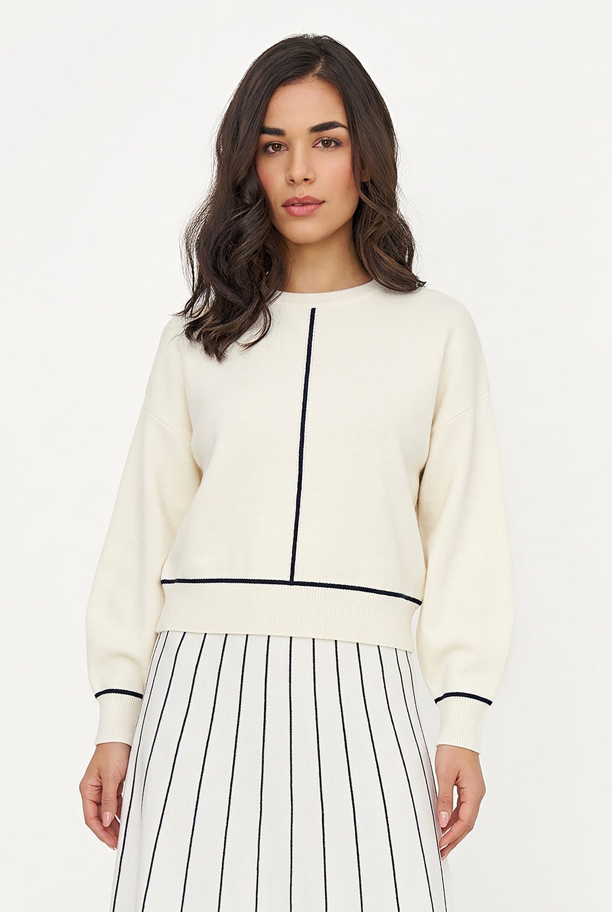 A woman modeling a cream-colored knit sweater with black trim details, paired with a black and white striped skirt, against a plain white studio background.