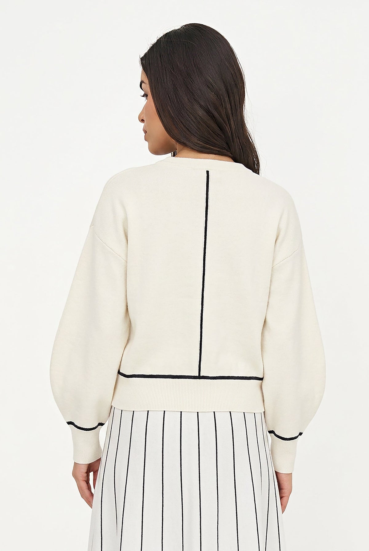A woman shown from the back wearing a cream-colored knit sweater with black contrast stitching details, paired with a white skirt featuring thin black vertical stripes, against a plain white studio background.