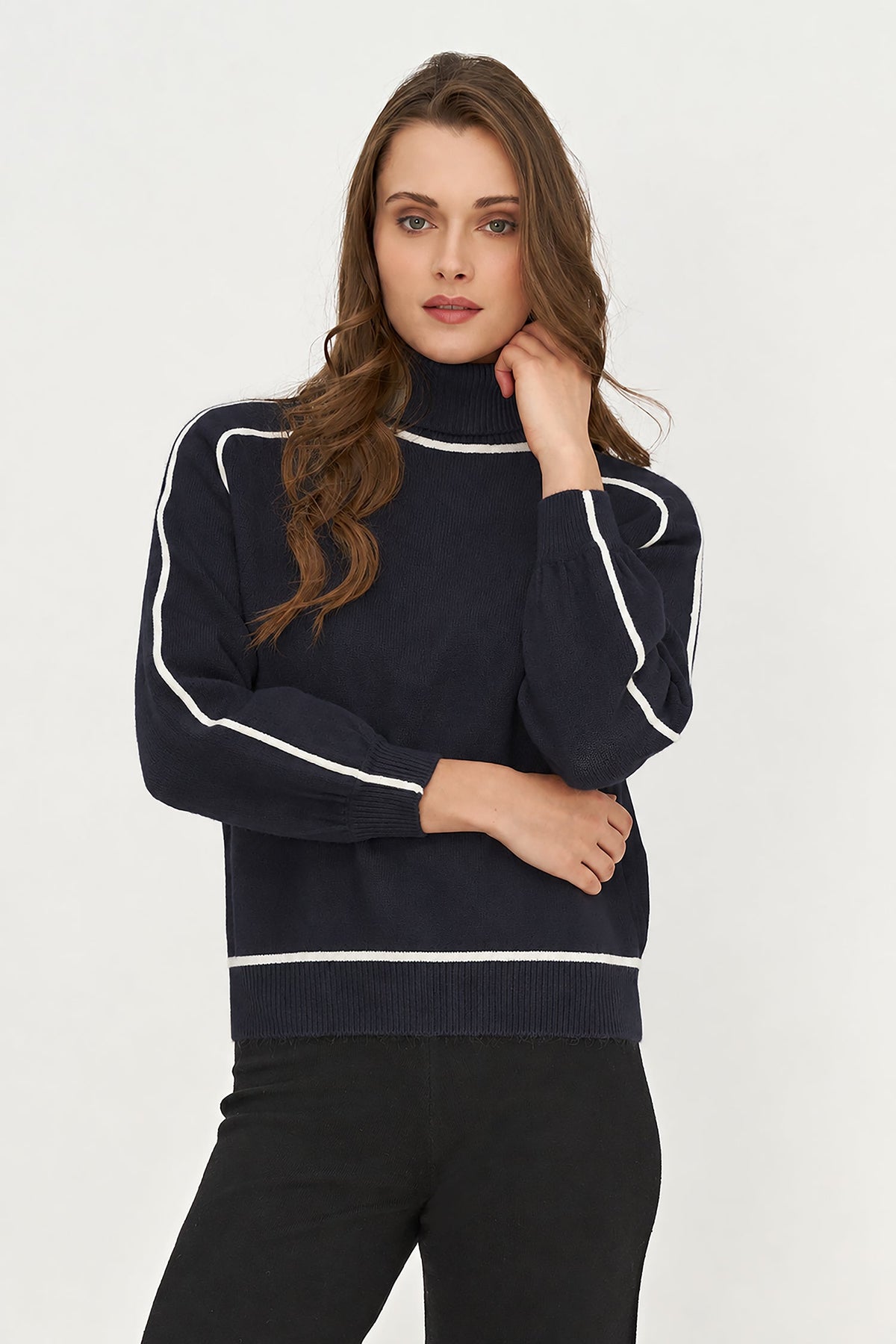 A woman modeling a navy blue mock neck sweater with white contrast piping details, paired with black pants, against a plain light gray background.
