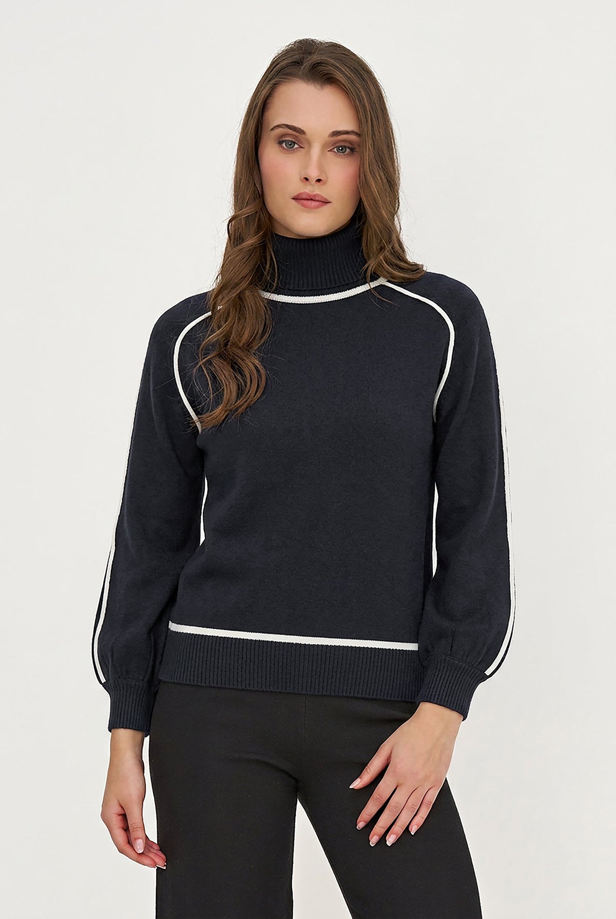 A woman modeling a dark navy blue mock-neck sweater with white contrast piping along the shoulders, sleeves, and hem, paired with black pants against a light gray studio background.