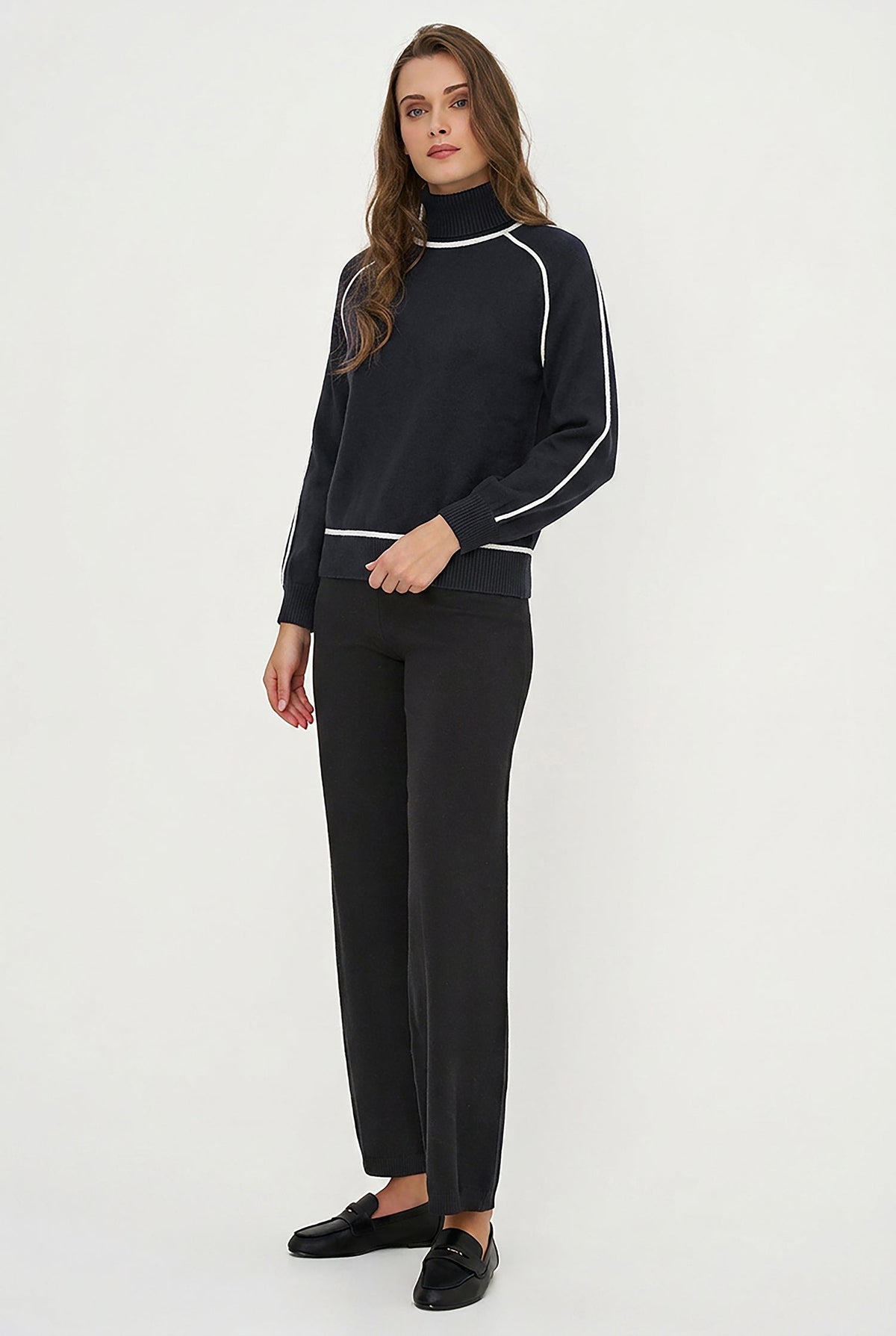 A woman modeling a black mock-neck long-sleeve top with white contrast stitching, paired with black trousers and black loafers, against a plain white studio background.