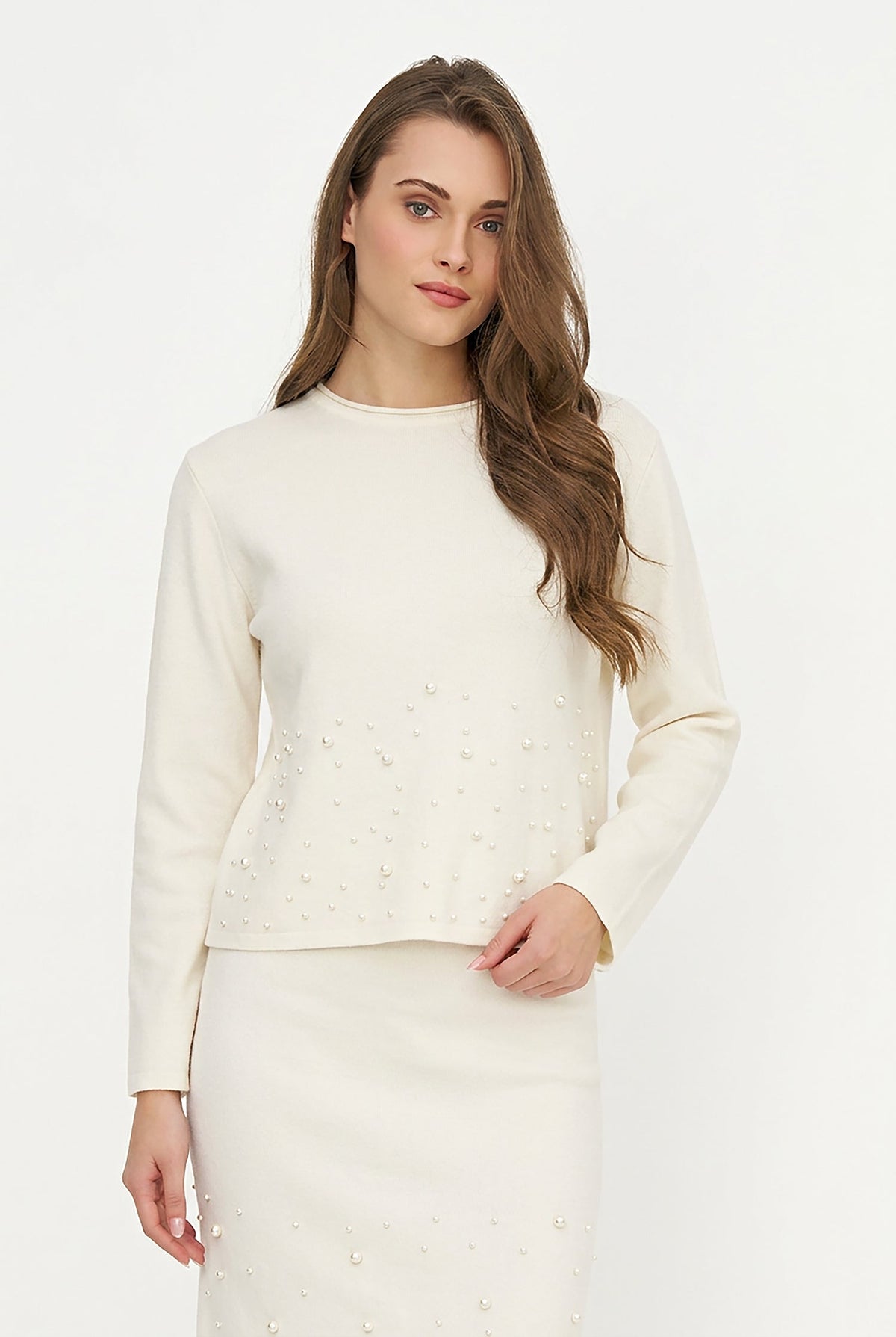 A woman modeling a cream-colored knit sweater and matching skirt set with pearl embellishments, set against a plain white studio background.
