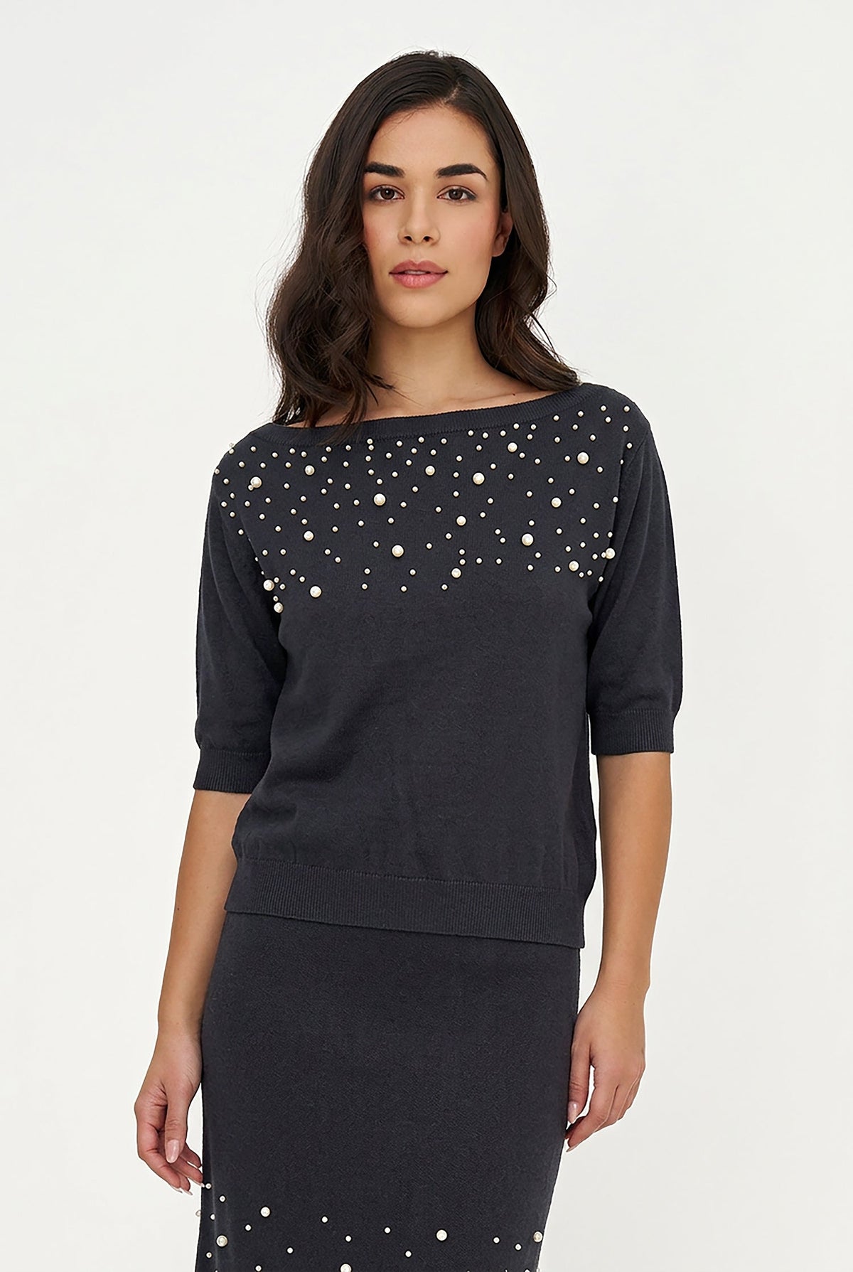 A woman modeling a dark charcoal gray knit dress with white pearl embellishments across the chest and along the skirt, set against a plain white studio background.