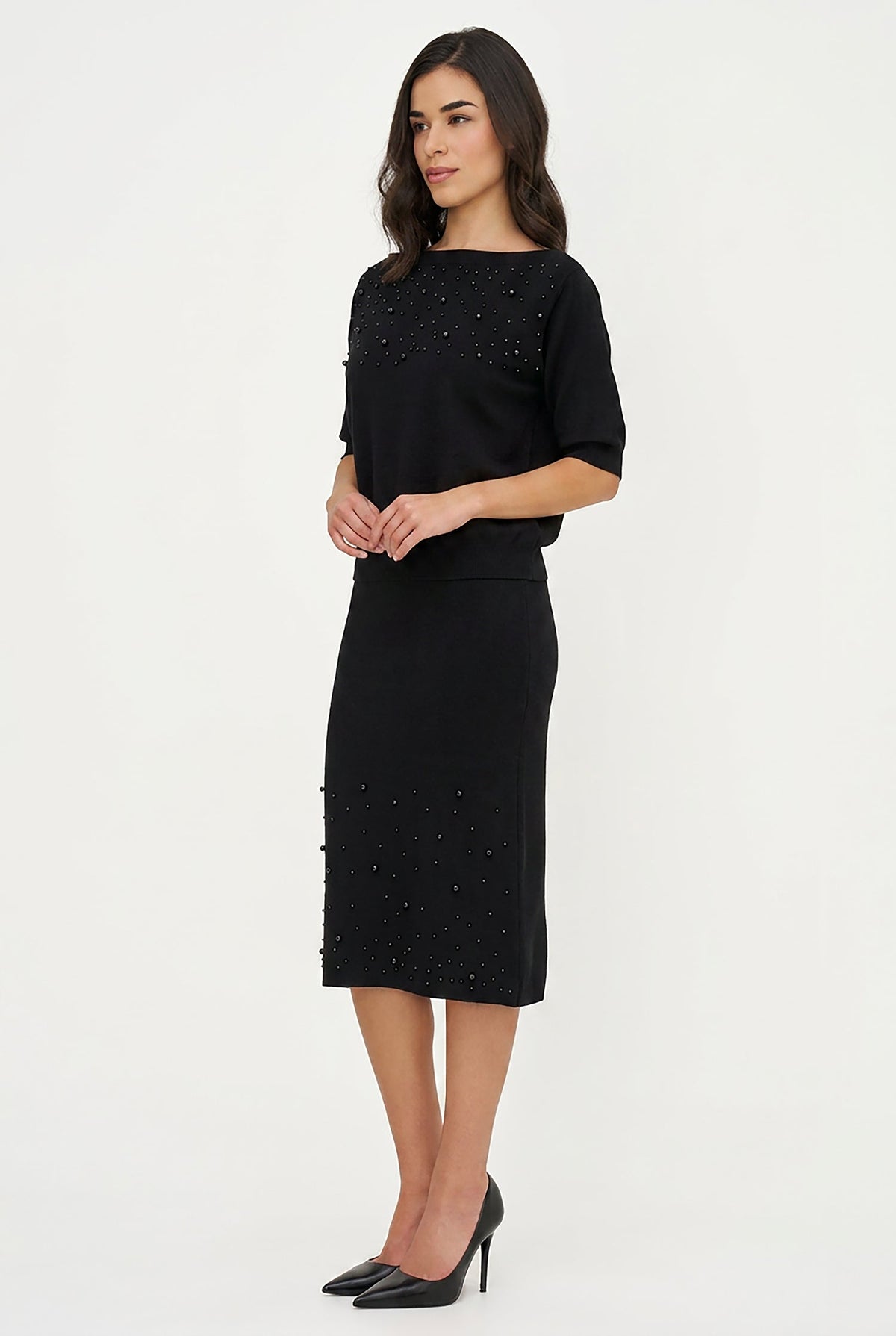 A woman modeling a black knee-length dress with elbow-length sleeves and delicate bead embellishments on the bodice and skirt, paired with black pointed-toe heels against a plain white studio background.
