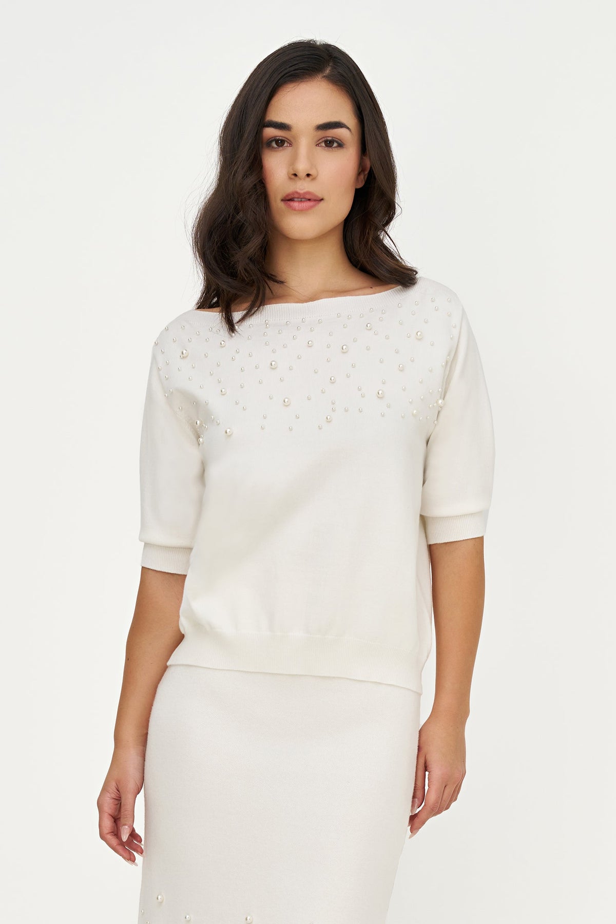A woman modeling a cream-colored knit top with short sleeves and delicate gold floral embroidery across the chest, paired with a matching cream skirt against a plain white studio background.
