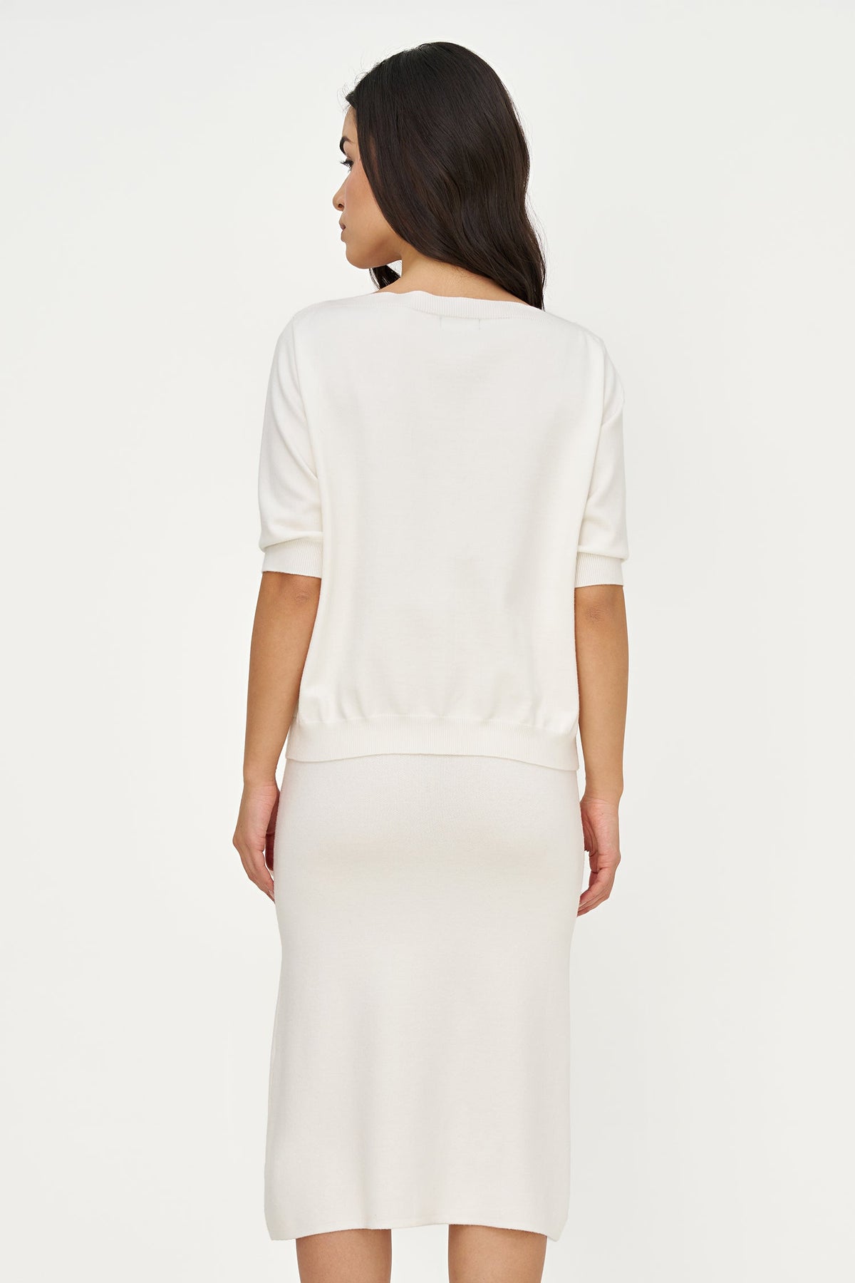 A woman shown from the back wearing a cream-colored knit dress with short sleeves and a knee-length skirt, set against a plain light gray studio background.