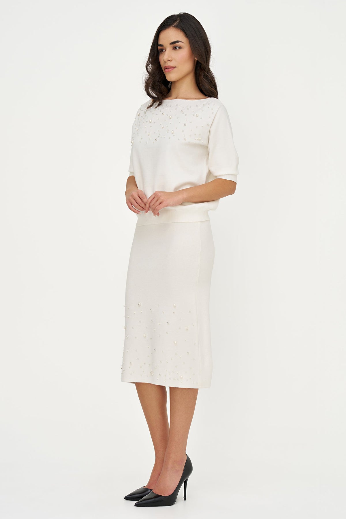 A woman modeling a cream-colored knit dress with short sleeves and a knee-length skirt, featuring delicate floral embroidery, paired with black pointed-toe heels against a plain white studio background.
