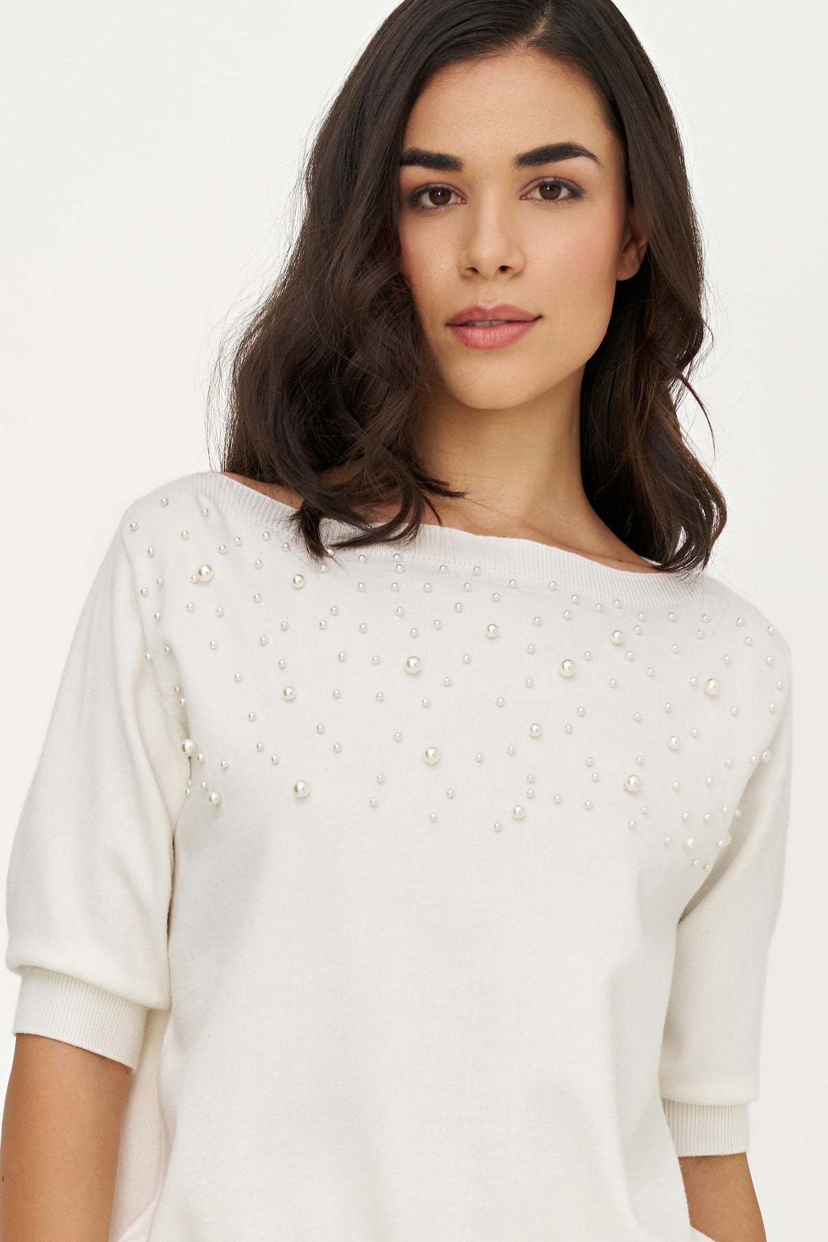 A woman wearing a cream-colored knit top with pearl embellishments across the chest, photographed against a plain light background.
